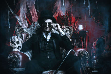 Halloween. Frightening gloomy man in a black tailcoat sits in an armchair with a skull in an old abandoned castle. The Dark Lord. Vampire man.の写真素材