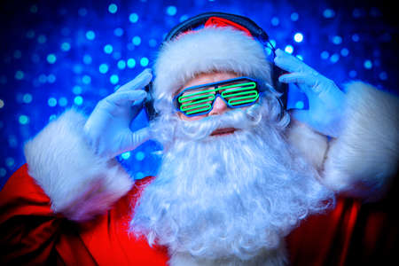 DJ Santa Claus in luminous glasses and headphones. Christmas songs and music. Disco lights in the background.の写真素材