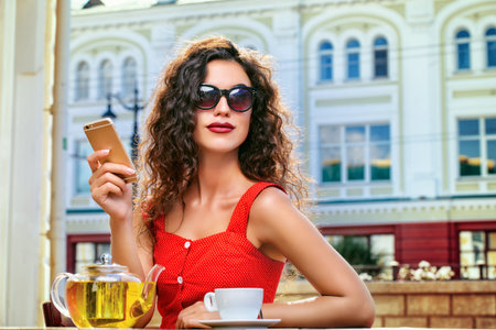 Charming brunette woman with beautiful smile using her smartphone while resting and drinking tea in a summer café.の写真素材