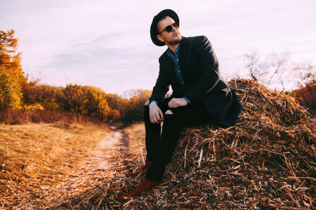 Autumn fashion. Handsome male model in black coat, a hat and sunglasses posing outdoor. Autumn nature.の写真素材