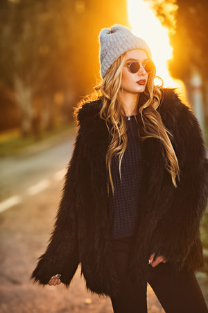 Autumn fashion shot. Trendy young woman in warm clothes posing on a highway on a sunset. Youth style.の写真素材