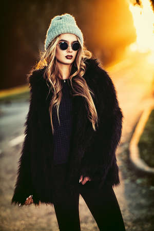 Autumn fashion shot. Trendy young woman in warm clothes posing on a highway on a sunset. Youth style.の写真素材