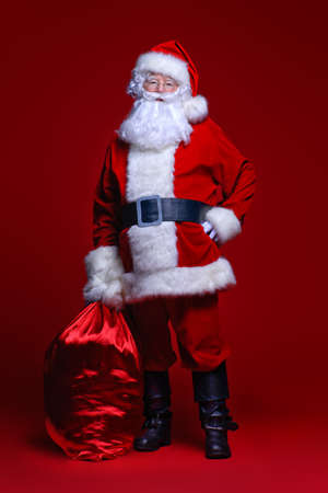 Christmas and New Year concept. Santa Claus comes with big bag of gifts. Full length portrait over red background.の写真素材