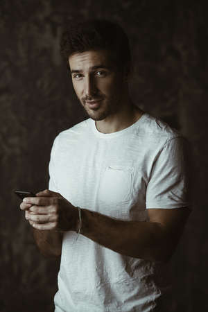 Handsome young man wearing white t-shirt  is holding his cell phone. Communications. Studio shot over dark background. Men's beauty and health.の写真素材