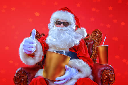 Merry Christmas and Happy New Year. Santa Claus sitting on his armchair and eating popcorn and drinking soda. Entertainment and cinema concept. Red background.の写真素材