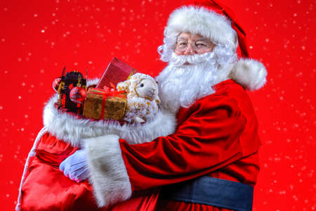 Christmas and New Year concept. Portrait of good old Santa Claus holding a sack with gifts and toys. Red background.の写真素材