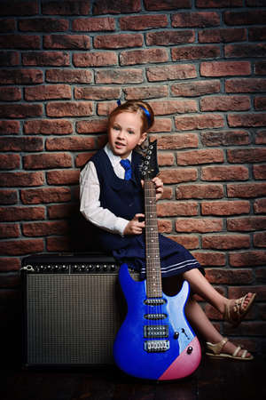 Modern little girl in school uniform posing with electric guitar over brick wall background. Rock star, rock music, pop music concept.の写真素材