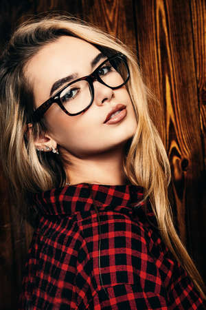 Portrait of a pretty blonde girl in checkered shirt and glasses standing by the wooden wall. Beauty, youth fashion. Student girl.の写真素材