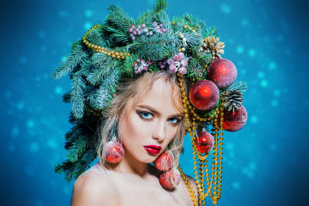 Portrait of a beautiful young woman with christmas tree in hairstyle decorated with beads and balls over blue background. Holiday make-up. Beauty, fashion.の写真素材