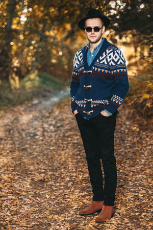 Full length portrait of a handsome man wearing cardigan and hat standing outdoor over autumn nature...