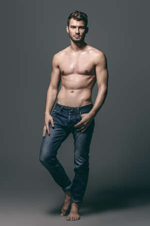 Full length portrait of a sexy young man in jeans and shirtless posing at studio. Gray background. Men's health. Beauty, jeans fashion.の写真素材