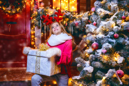 Pretty child girl is sitting on the porch of a house decorated for Christmas and holding a gift. Time for miracles. の写真素材