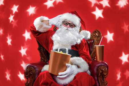 Merry Christmas and Happy New Year. Santa Claus sitting on his armchair and eating popcorn and drinking soda. Entertainment and cinema concept. Red background.の写真素材