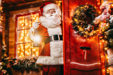 Christmas concept. Portrait of a fairytale Santa Claus peek out from behind the door with lantern and wonders. Beautiful house decorated for Christmas. Time of miracles.の写真素材
