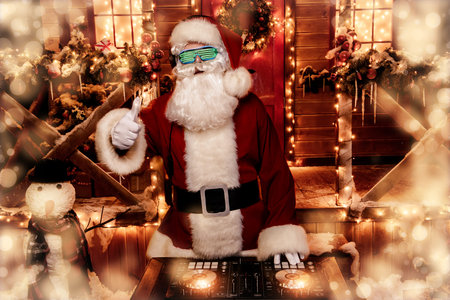 DJ Santa Claus in luminous glasses and headphones holds a party near his house decorated with...