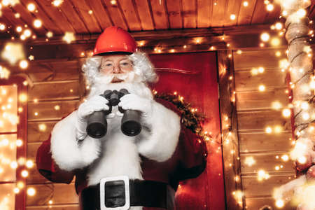 Santa Claus is a builder. House of Santa Claus. Portrait of Santa Claus in a helmet on his head standing near his decorated house and looks through binoculars. Christmas and New Year concept.の写真素材