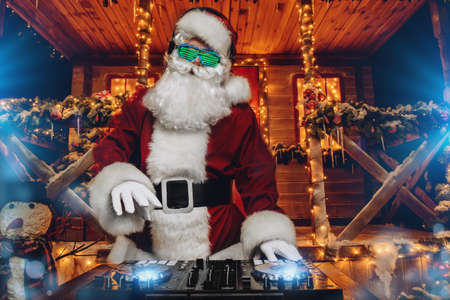 DJ Santa Claus in luminous glasses and headphones holds a party near his house decorated with lights. Christmas songs and music. の写真素材