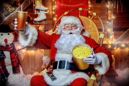 Santa Claus sitting on his armchair eating popcorn, drinking soda and watching a Christmas movie. Entertainment and cinema concept. Merry Christmas and Happy New Year.の写真素材