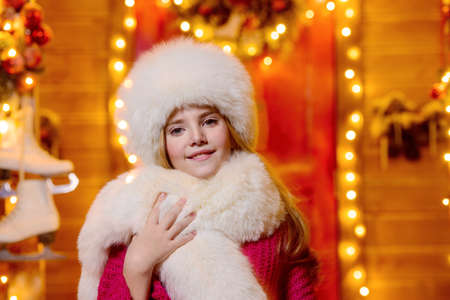 Portrait of a beautiful nine-year-old girl in a fur hat and scarf near her house decorated for Christmas.の写真素材