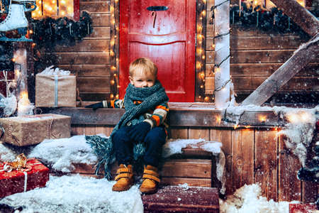 Cute little boy in winter clothes sits on the porch of the magic house of Santa Claus. Miracle time. Merry Christmas and Happy New Year.の写真素材