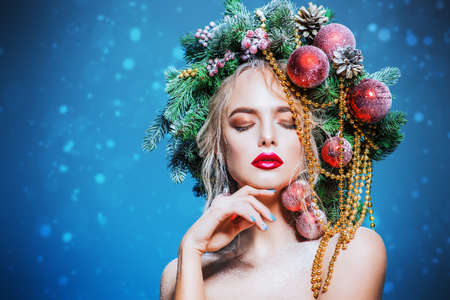 Portrait of a beautiful young woman with christmas tree in hairstyle decorated with beads and balls over blue background. Holiday make-up. Beauty, fashion.の写真素材