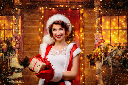 Smiling Santa girl holds a gift box in her hands and poses near the house of Santa, decorated with festive lights. Christmas and New Year concept.の写真素材