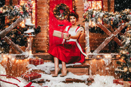 Smiling Santa girl holds a gift box in her hands and poses near the house of Santa, decorated with festive lights. Christmas and New Year concept.の写真素材