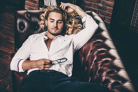 Handsome sexy young man in white shirt lying on a sofa. Men's beauty, fashion. Hair, hairstyle.の写真素材