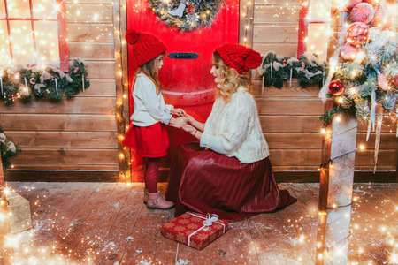 Happy mother and daughter in beautiful warm clothes stand on the porch of the house, decorated for Christmas. Seasonal winter fashion. Merry Christmas and Happy New Year.の写真素材