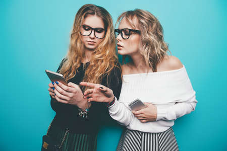 Two pretty girls using their smartphones and  posing in studio over blue background. Modern lifestyle. Studio shot.の写真素材