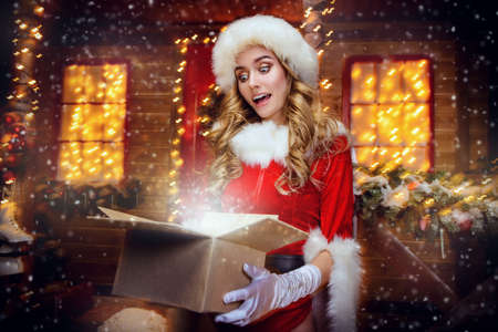 Santa girl in red body suit opens a gift box and surprises, near the house of Santa, decorated with festive lights. Christmas and New Year concept.の写真素材