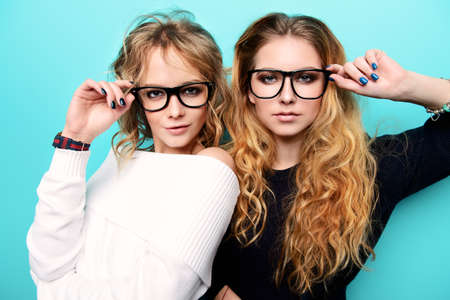 Two pretty girls in glasses are posing in studio over blue background. Beauty, fashion. Optics, eyewear.の写真素材