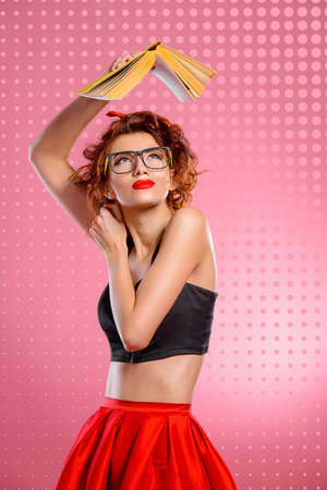 Funny pretty girl in glasses is reading a book over pink background. Pin-up retro style.の写真素材