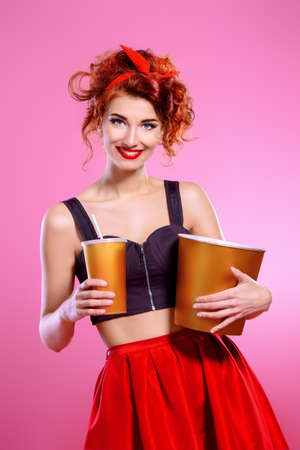 Pretty emotional young woman eating popcorn and drinking soda over pink background. Pin-up style. Entertainment and cinema concept.の写真素材