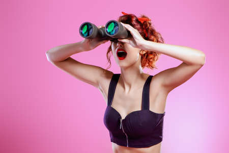 Pretty red-haired girl looks through binoculars at something over pink background. Pin-up style. の写真素材
