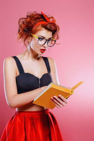 Smart pretty girl in glasses is reading a book over pink background. Pin-up retro style.の写真素材