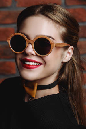 Stylish girl in black dress and round sunglasses posing by the brick wall. Modern youth fashion.の写真素材
