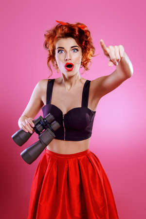 Pretty red-haired girl with binoculars, looks at something over pink background. Pin-up style. の写真素材