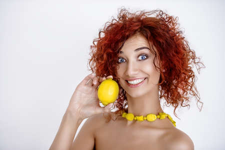 Beauty, natural cosmetics and hair concept. Beautiful red-haired young woman in yellow beads holding a lemon. White background.の写真素材