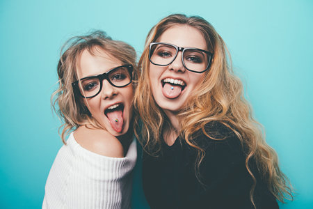 Two cheerful pretty girls in glasses stick out their tongue. Blue background. Beauty, fashion....