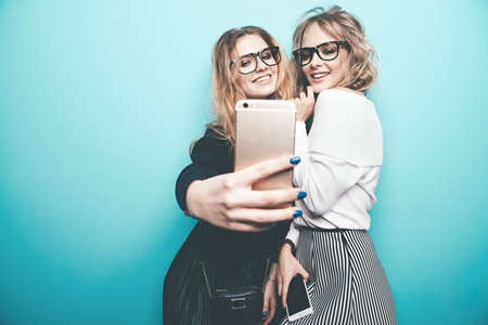 Two pretty girls are making selfie. Modern lifestyle. Studio shot over blue background.の写真素材