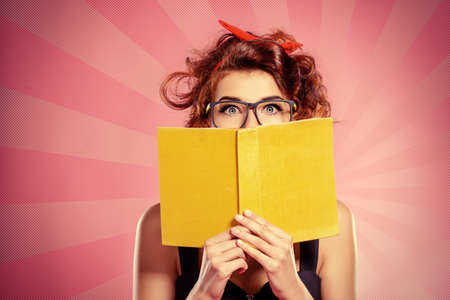 Smart pretty girl in glasses is reading a book over pink background. Pin-up retro style.の写真素材