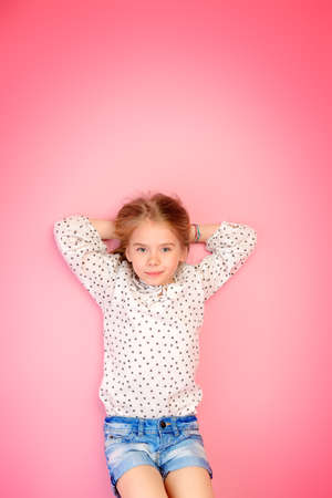 Happy childhood. Cute eight year old girl wearing summer jeans shorts and blouse lying on a pink floor. Studio shot. の写真素材
