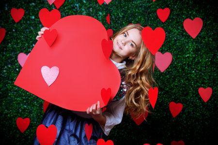 Happy pre-teen girl holding red heart surrounded by little hearts over lawny background. First love....