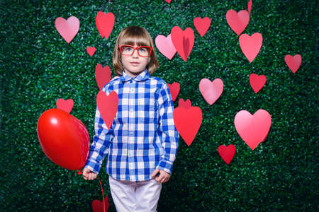 Ð¡ute child boy holding heart shaped balloon, surrounded by hearts over lawny background. First love. Valentine's Day. の写真素材
