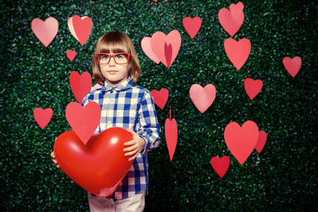 Ð¡ute child boy holding heart shaped balloon, surrounded by hearts over lawny background. First love. Valentine's Day. の写真素材