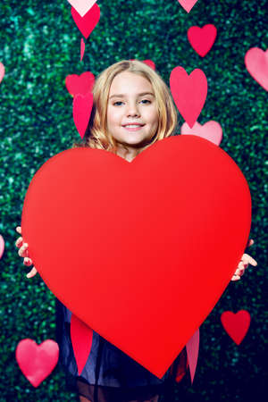 Happy pre-teen girl holding red heart surrounded by little hearts over lawny background. First love. Valentine's Day. の写真素材