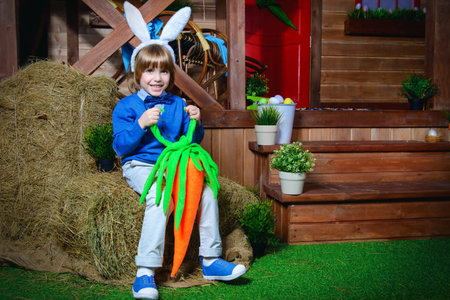 Funny little boy with bunny ears and huge carrot sitting on a haystack in the yard near the wooden...