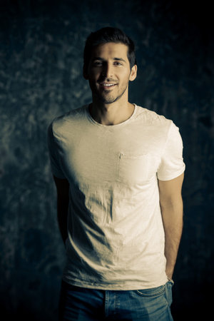 Portrait of a handsome smiling young man wearing white t-shirt. Studio shot over dark background....