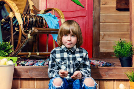 Happy little boy sitting on the porch near the wooden house. Easter holiday. Rural style, easter decoration.の写真素材